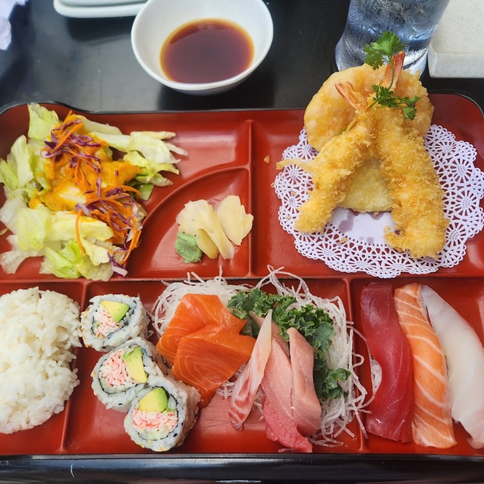 Tsunami Sushi bento box with tempura shrimp, California rolls, sashimi, nigiri, rice, and salad.