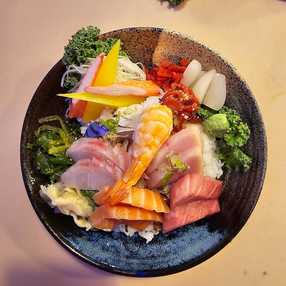 A colorful Tsunami Sushi chirashi bowl with various sashimi, shrimp, and vegetables over rice.