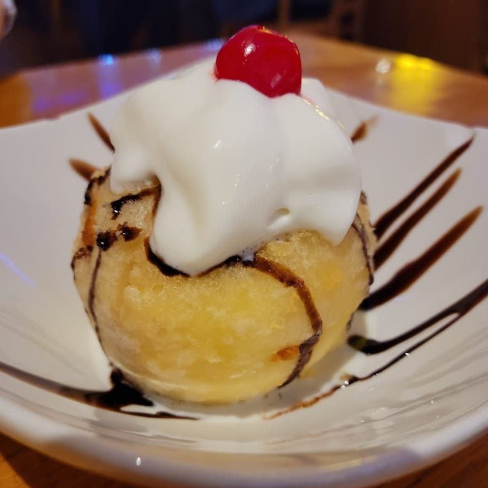 Fried ice cream dessert with whipped cream, chocolate drizzle, and a cherry on a white plate.